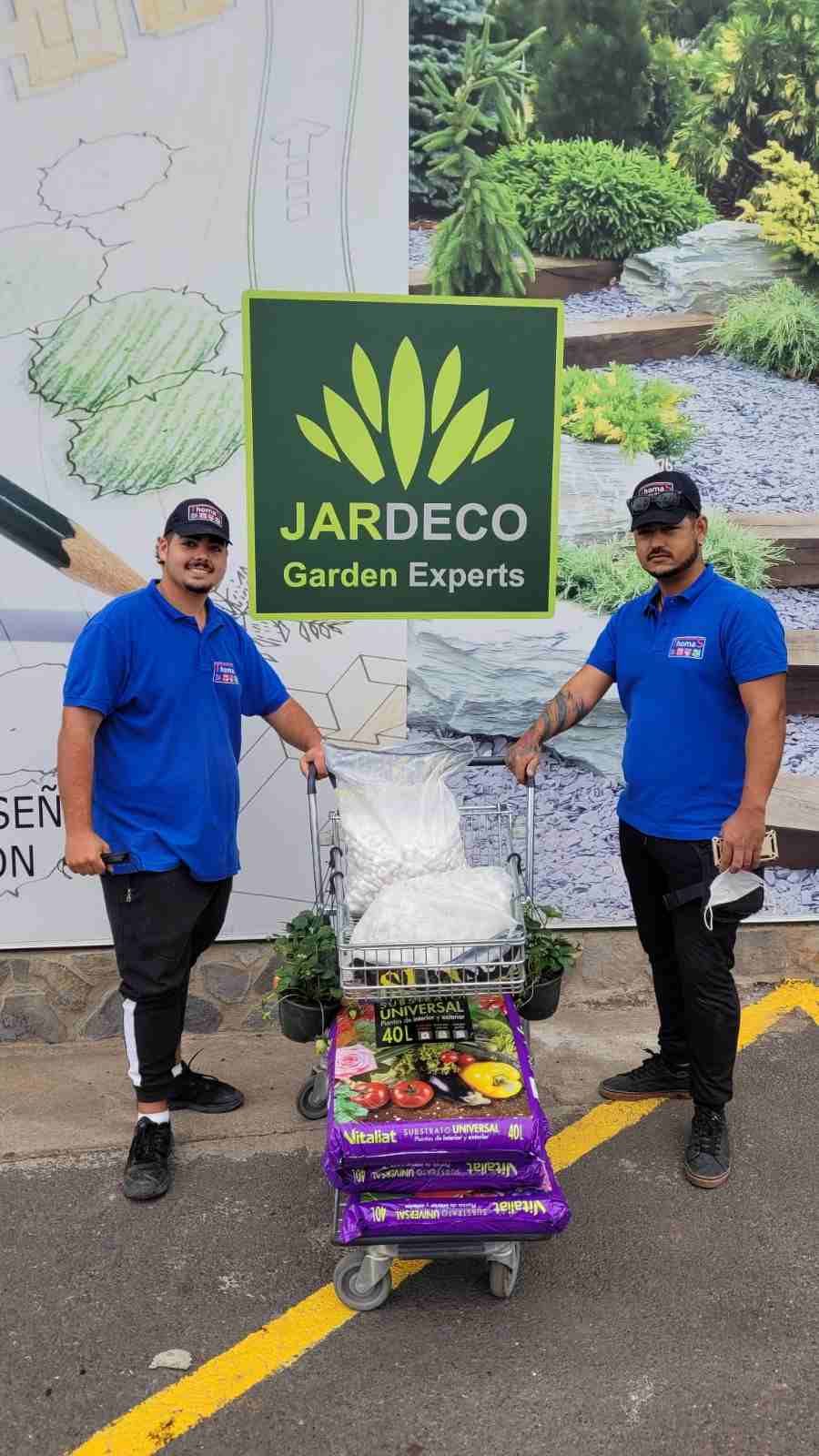 Dos jardineros con camisas azules empujan un carrito con plantas frente a un letrero de Jardeco Garden Experts.