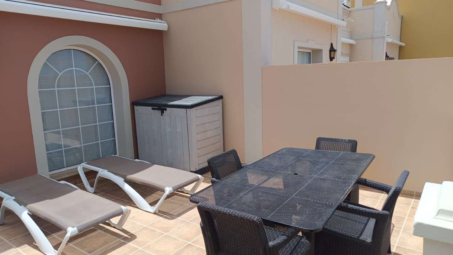 Patio con tumbonas y una mesa de comedor de mimbre oscuro junto a una pared de color melocotón.