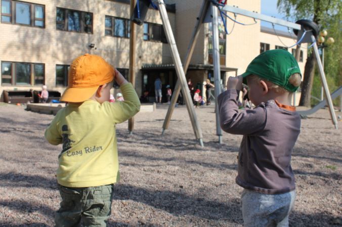 Kaksi värikkäitä hattuja käyttävää lasta seisoo leikkikentällä keinujen ja rakennuksen lähellä