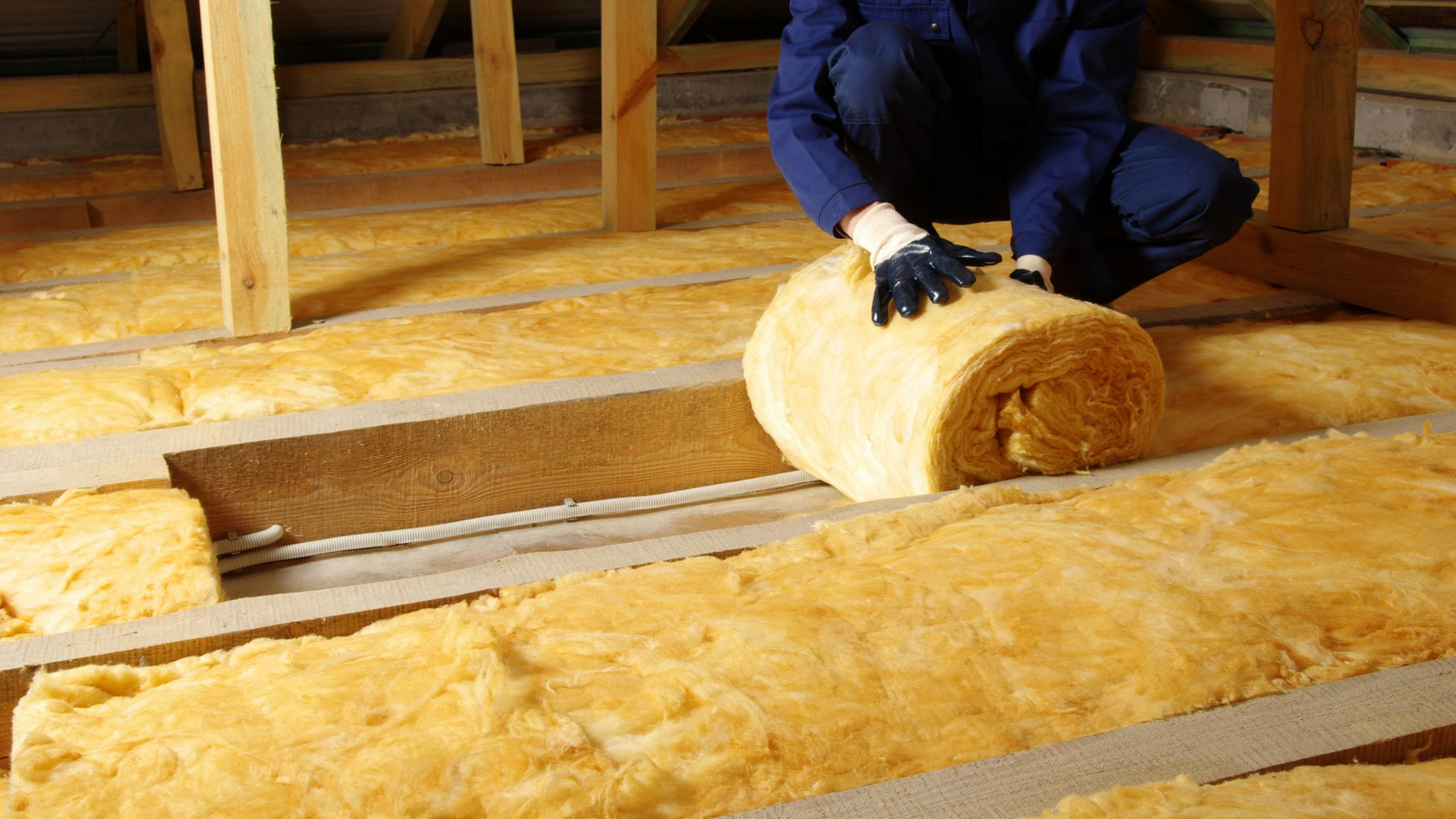Une personne installe de l'isolant dans un grenier, déroulant de l'isolant jaune entre des poutres en bois.