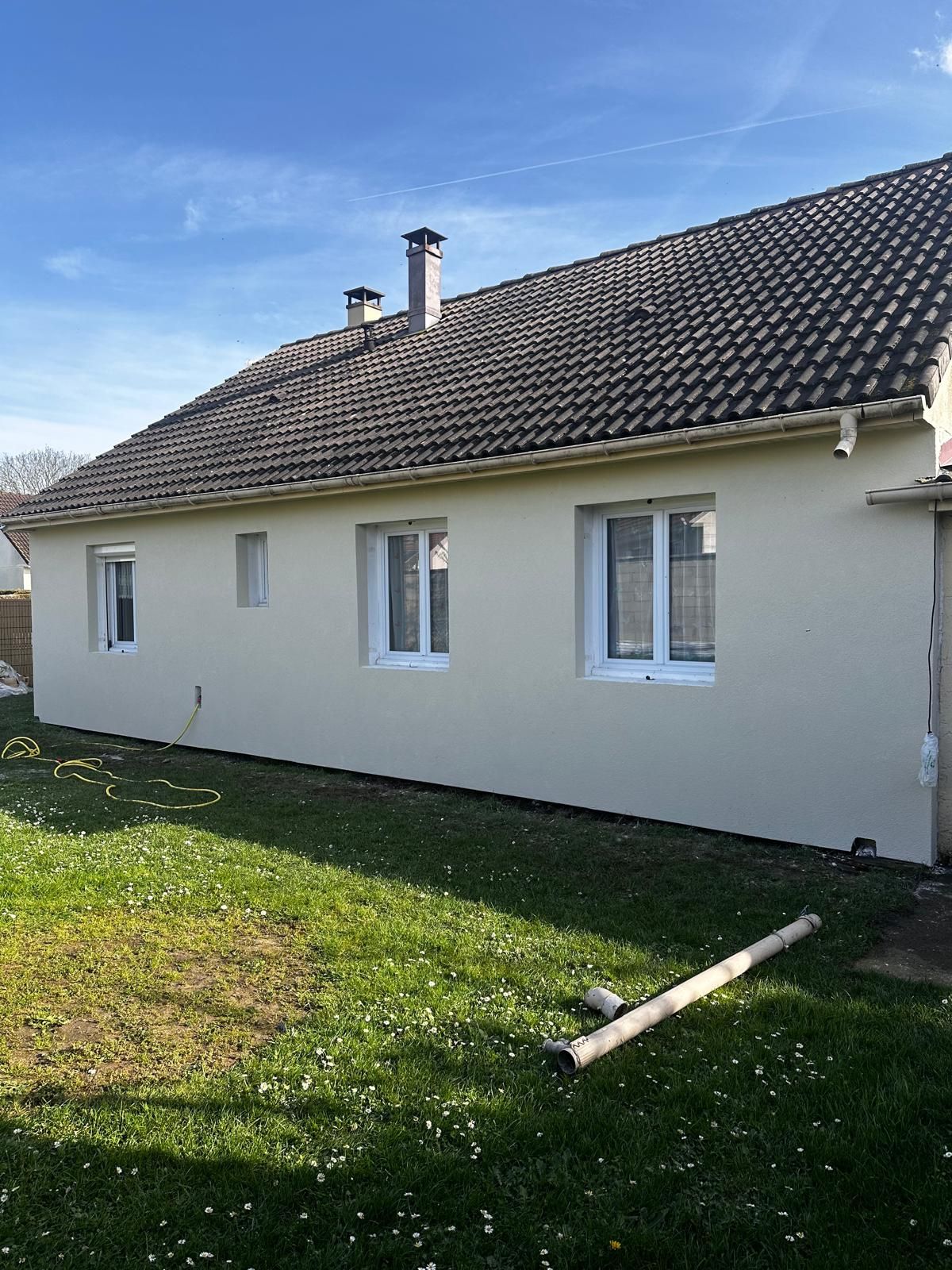 Maison beige aux fenêtres à cadre blanc, toit de tuiles foncées et pelouse verte par une journée ensoleillée.