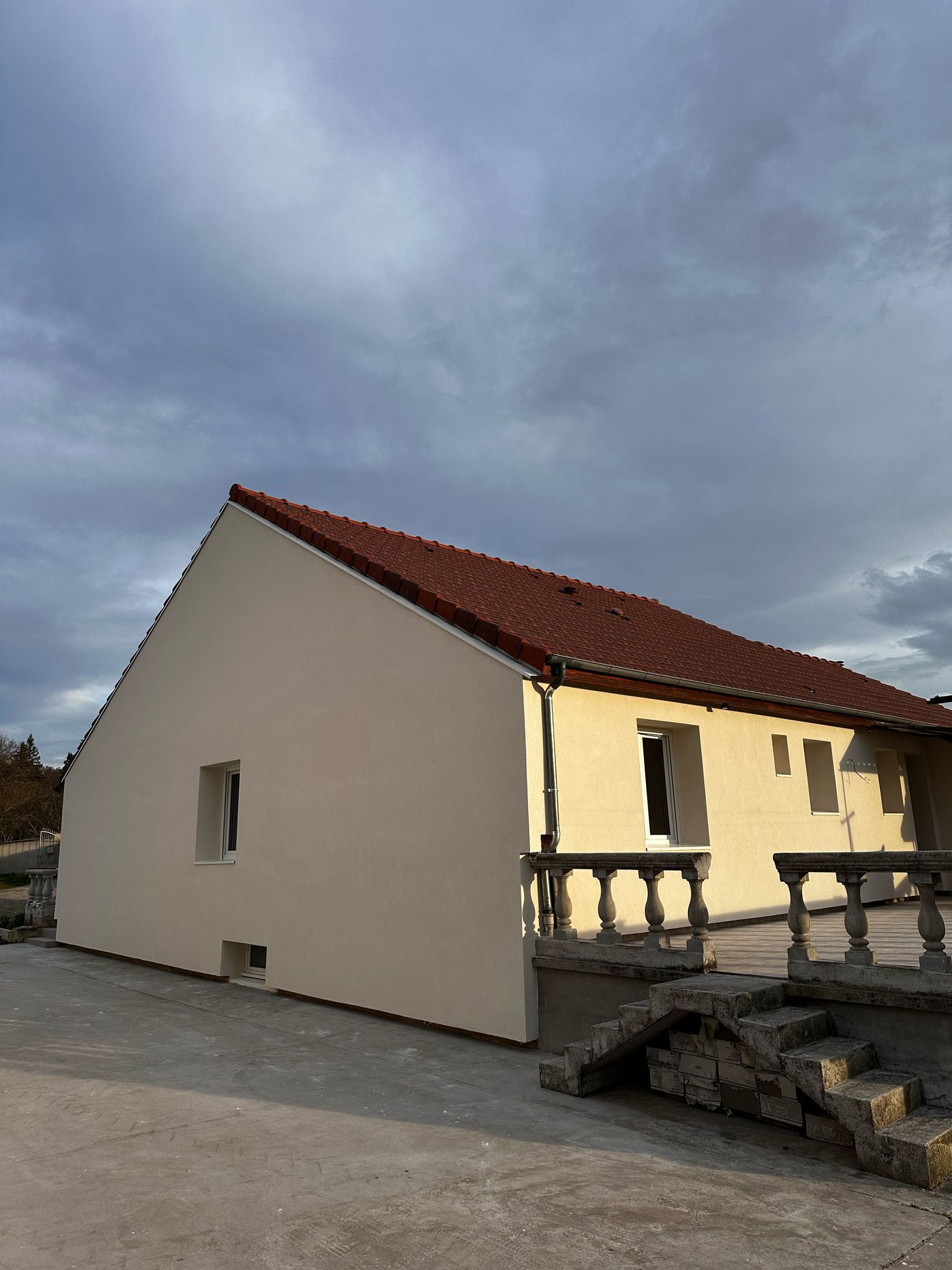 Maison beige au toit de tuiles rouges sous un ciel nuageux. La maison possède une petite fenêtre et un balcon.
