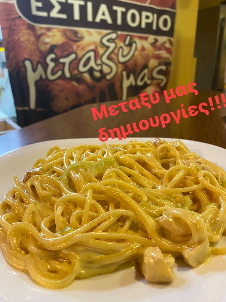 Plate of creamy pasta in a restaurant, with Greek menu sign in the background and red text overlay stating “Μεταξύ μας δημιουργίες!!”