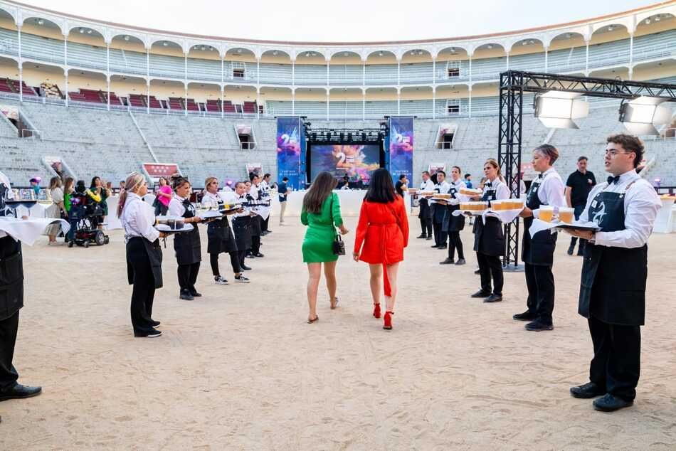Dos mujeres con vestidos coloridos caminan hacia un escenario en una plaza de toros, flanqueadas por camareros que sostienen bandejas de comida.