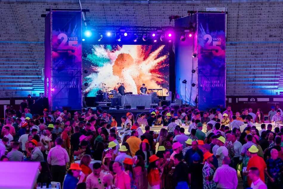 Multitud en un concierto al aire libre bien iluminado con un DJ en el escenario frente a una pantalla grande con gráficos coloridos.