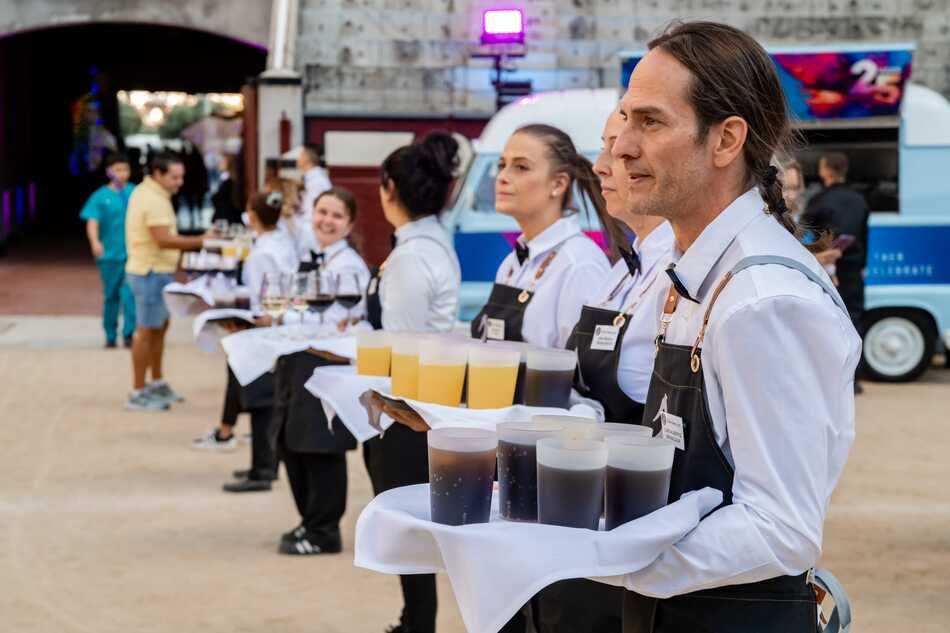 Camareros con bandejas de bebidas en un evento al aire libre. Llevan delantales negros y camisas blancas. Se ve un food truck al fondo.
