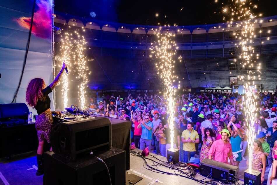 DJ actuando en el escenario con pirotecnia en un concierto al aire libre. El público aplaude, iluminado por las luces del escenario.