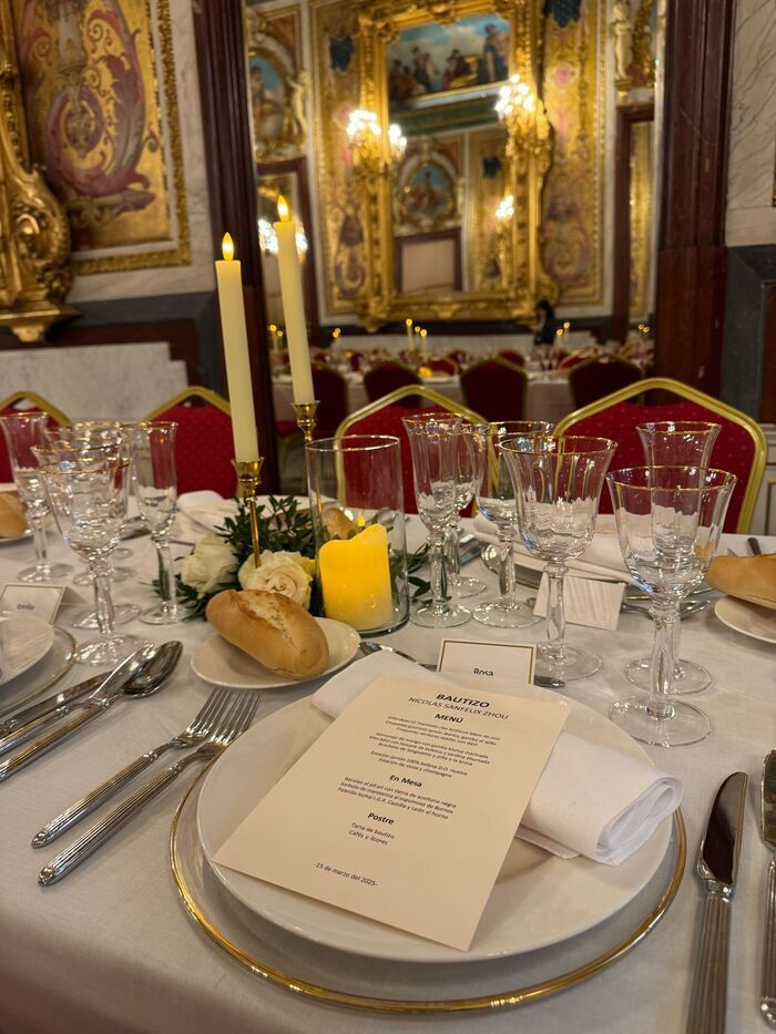 Mesa de comedor formal con mantel blanco, platos con borde dorado, copas de cristal y menú. A la luz de las velas, en una habitación decorada con espejos.