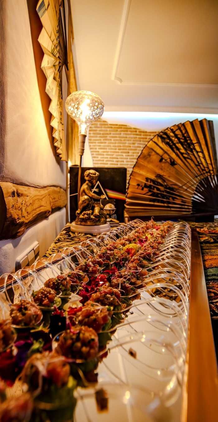 Mesa preparada para evento, decorada con comida y luces. Estatua de Buda y abanico decorativo al fondo.