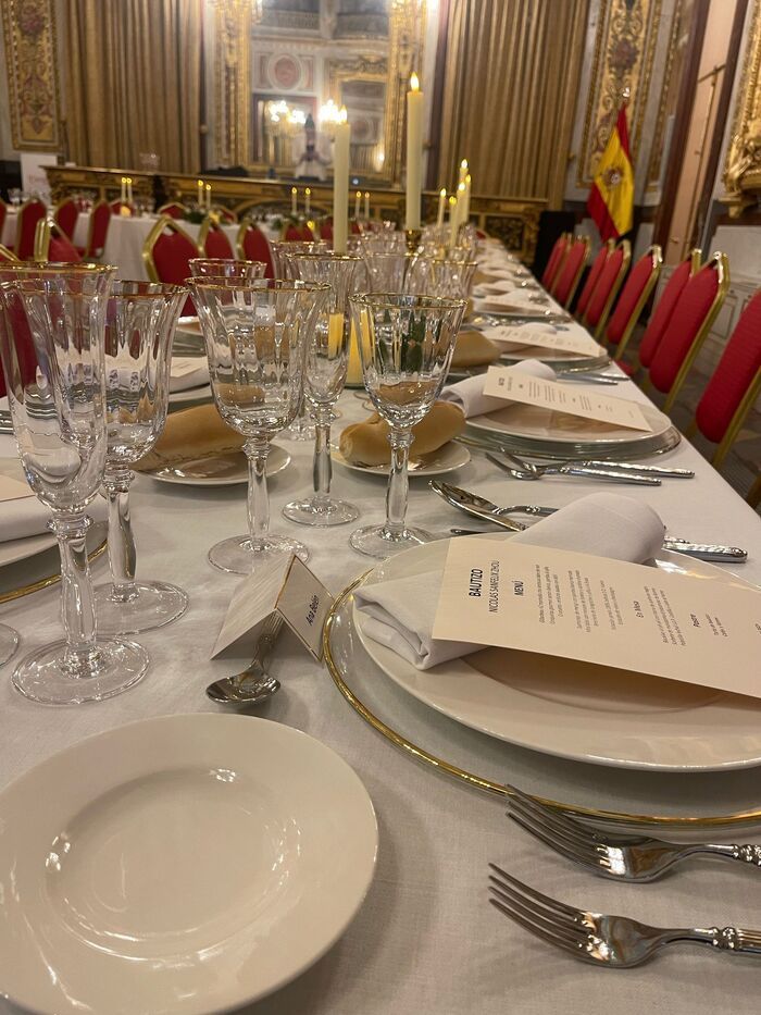 Mesa de comedor formal dispuesta para banquete, con mantel blanco, cubertería, cristalería y tarjetas de menú; bandera española de fondo.