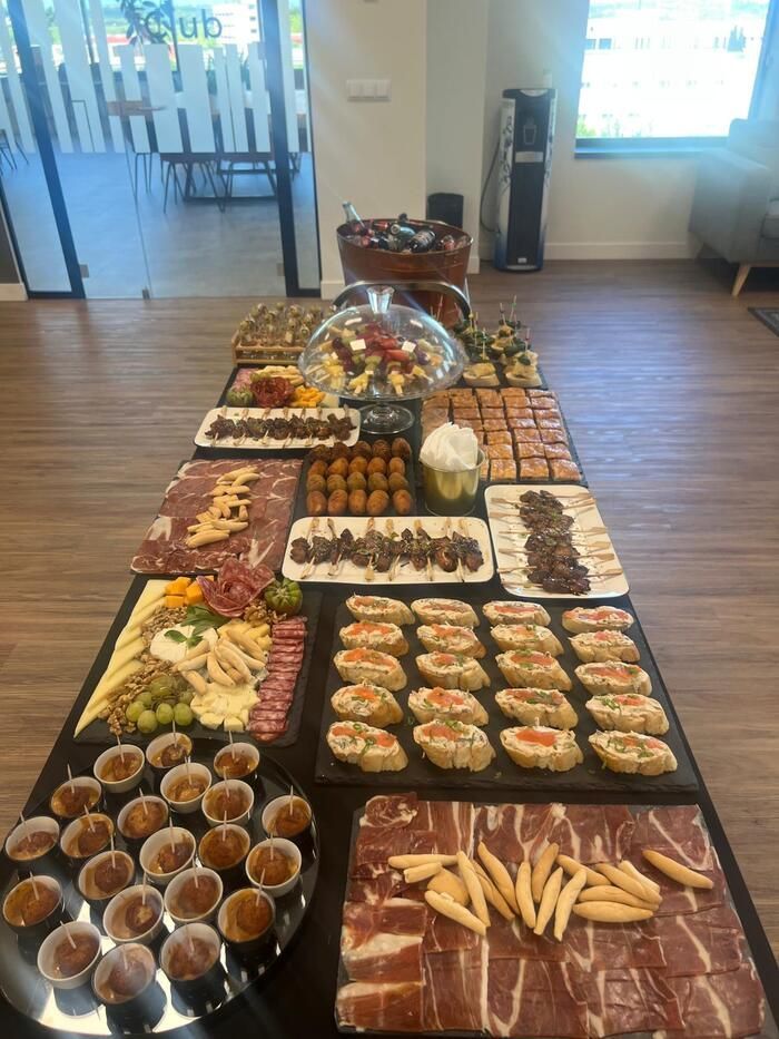 Una mesa larga repleta de aperitivos variados, incluyendo carnes, quesos y canapés, dispuesta para una reunión en una sala bien iluminada.