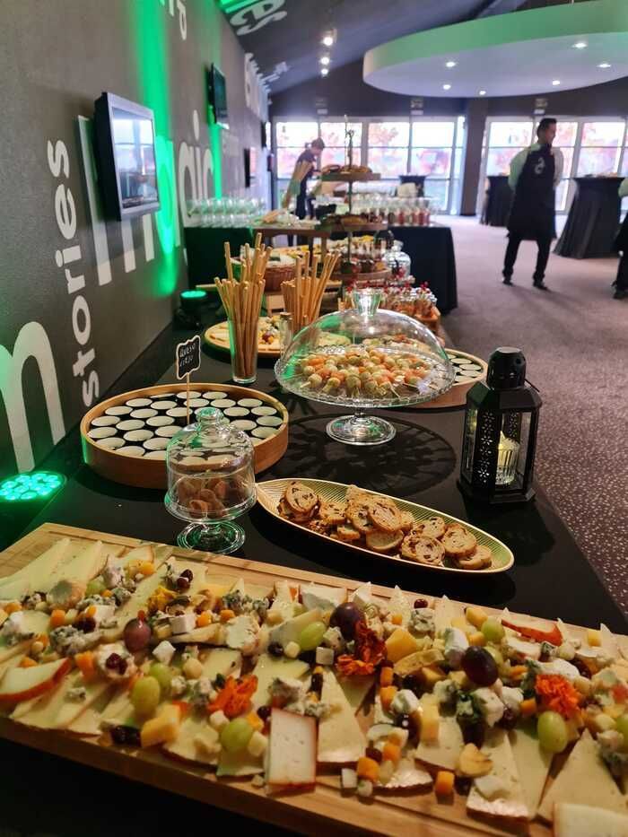 Mesa de buffet con queso, galletas y bocadillos en un evento. Una persona con delantal está de pie al fondo.