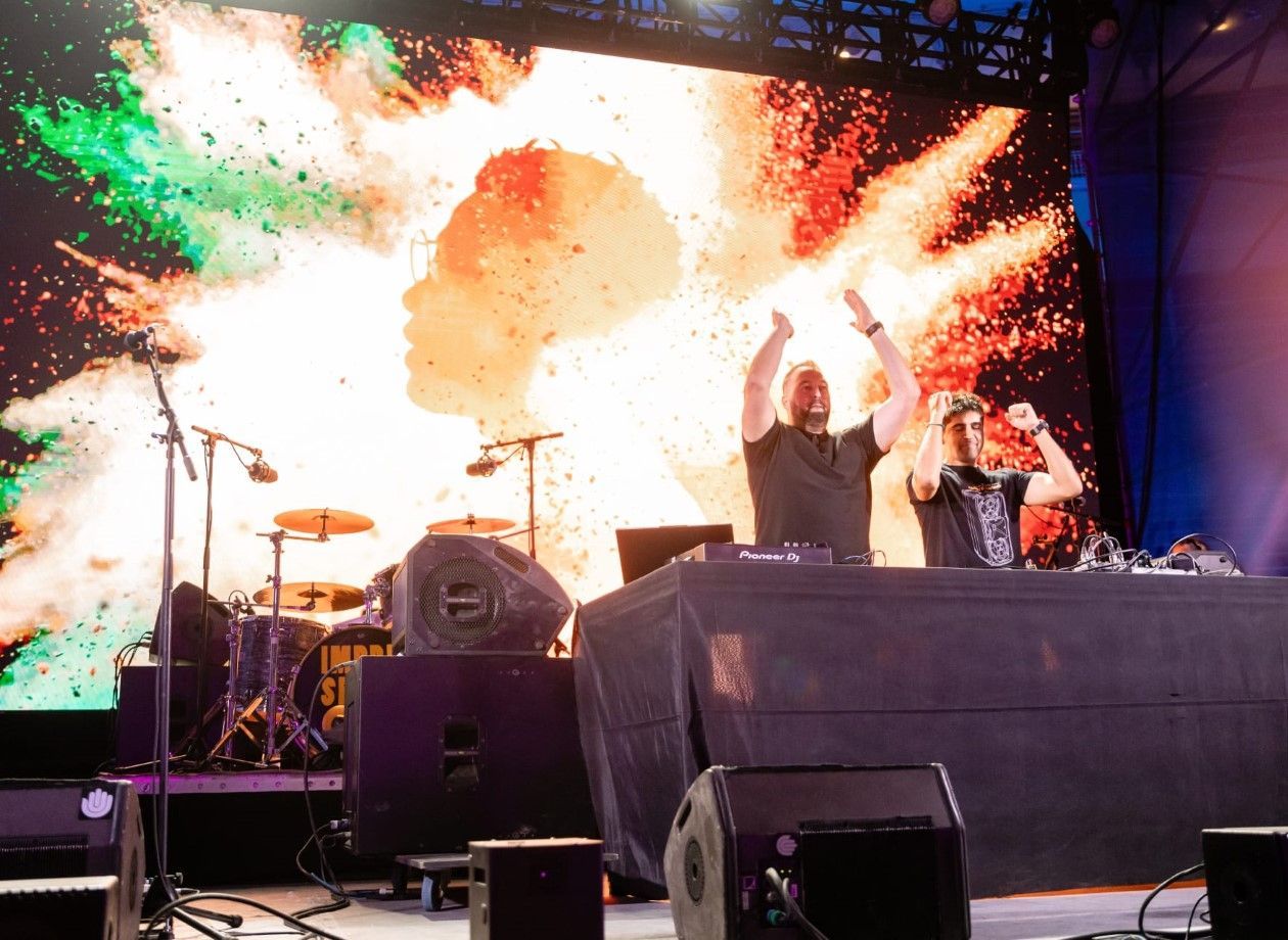 Dos DJs en un escenario con los brazos en alto. Una vibrante explosión de colores y una silueta de fondo detrás.