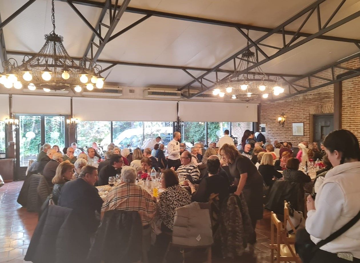 Un gran grupo de personas sentadas a mesas en un espacio para eventos con una cálida iluminación. Lámparas de araña cuelgan del techo alto, con un altavoz al frente.
