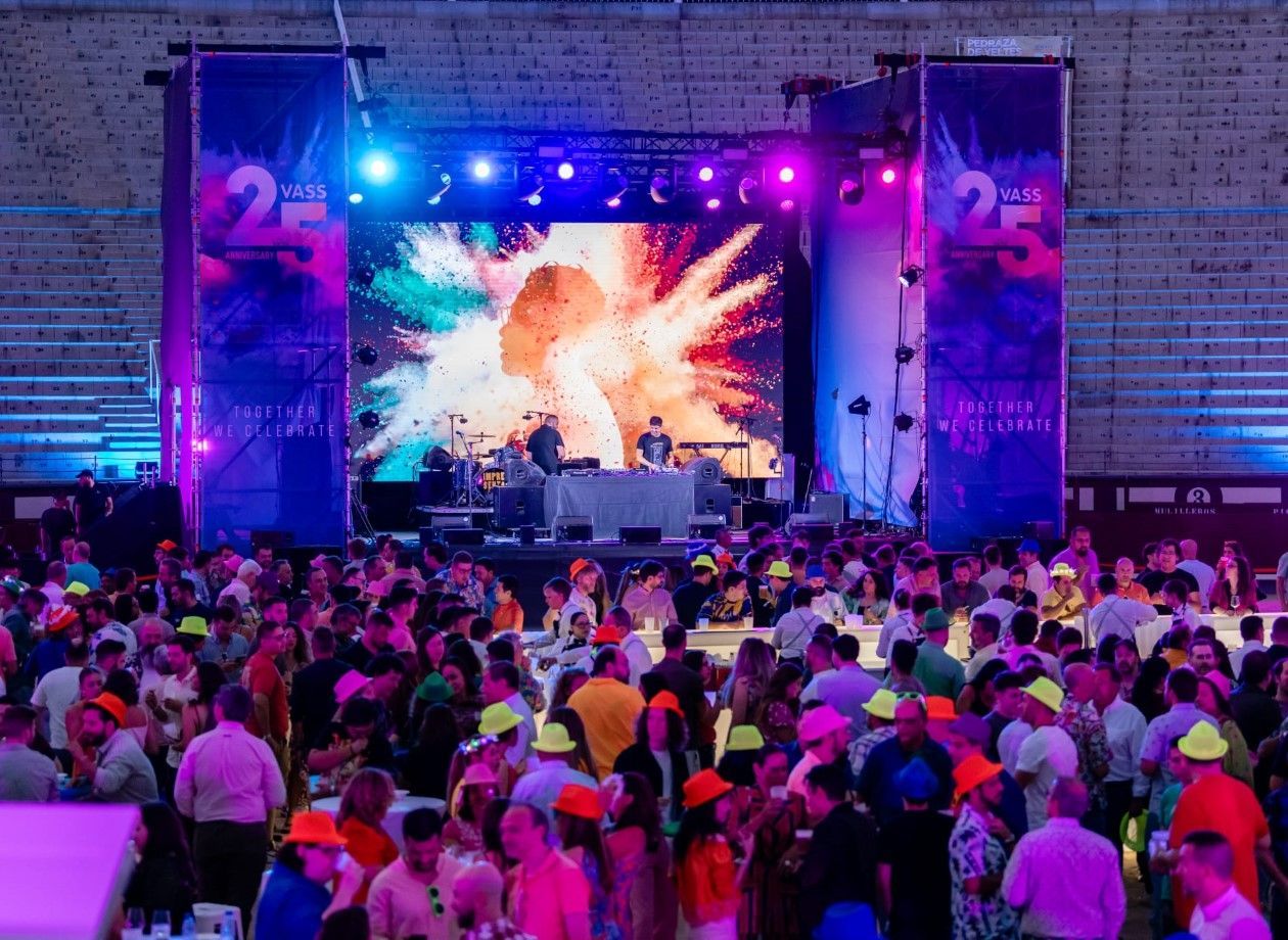 Una gran multitud asiste a un concierto en un escenario con luces de colores y una pantalla de video. La gente lleva sombreros llamativos.