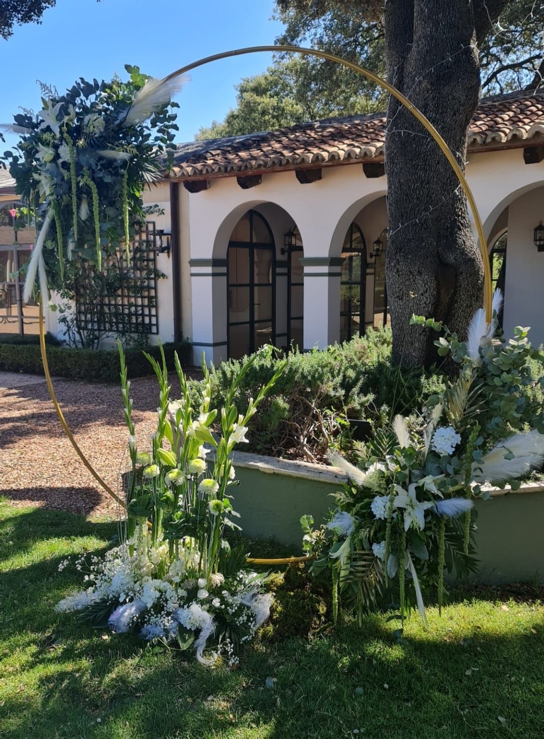 Un arco de boda circular dorado decorado con flores blancas se encuentra sobre un césped verde con un edificio al fondo.