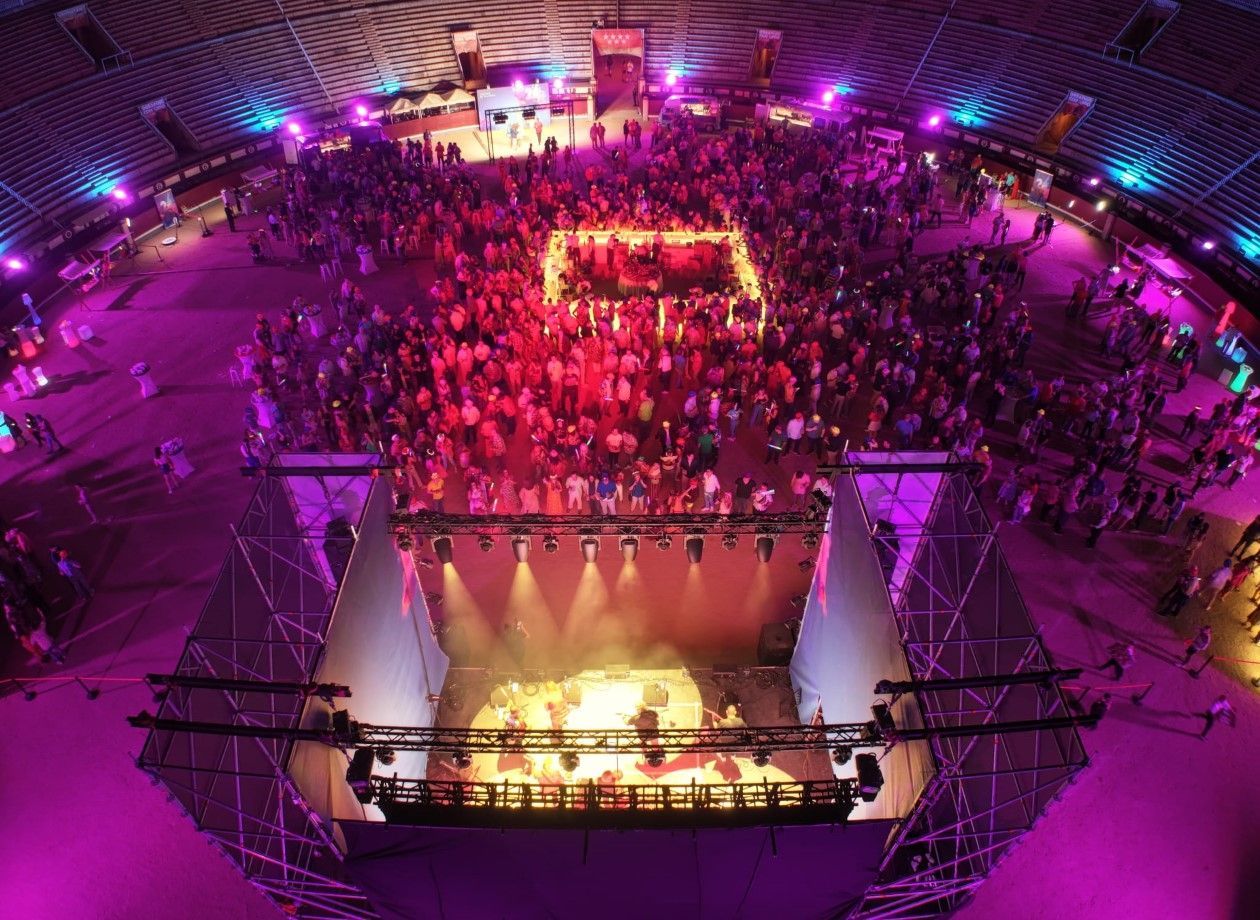 Gran sala de conciertos con escenario iluminado por focos y una multitud. Fotografía cenital con vibrantes luces rosas y moradas.