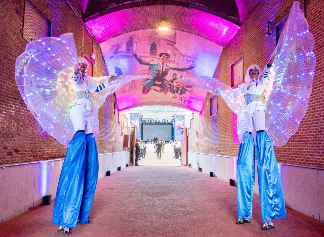 Dos artistas en zancos con alas brillantes se encuentran en un arco de ladrillo. Llevan trajes blancos con detalles azules y miran hacia un espacio de eventos distante.