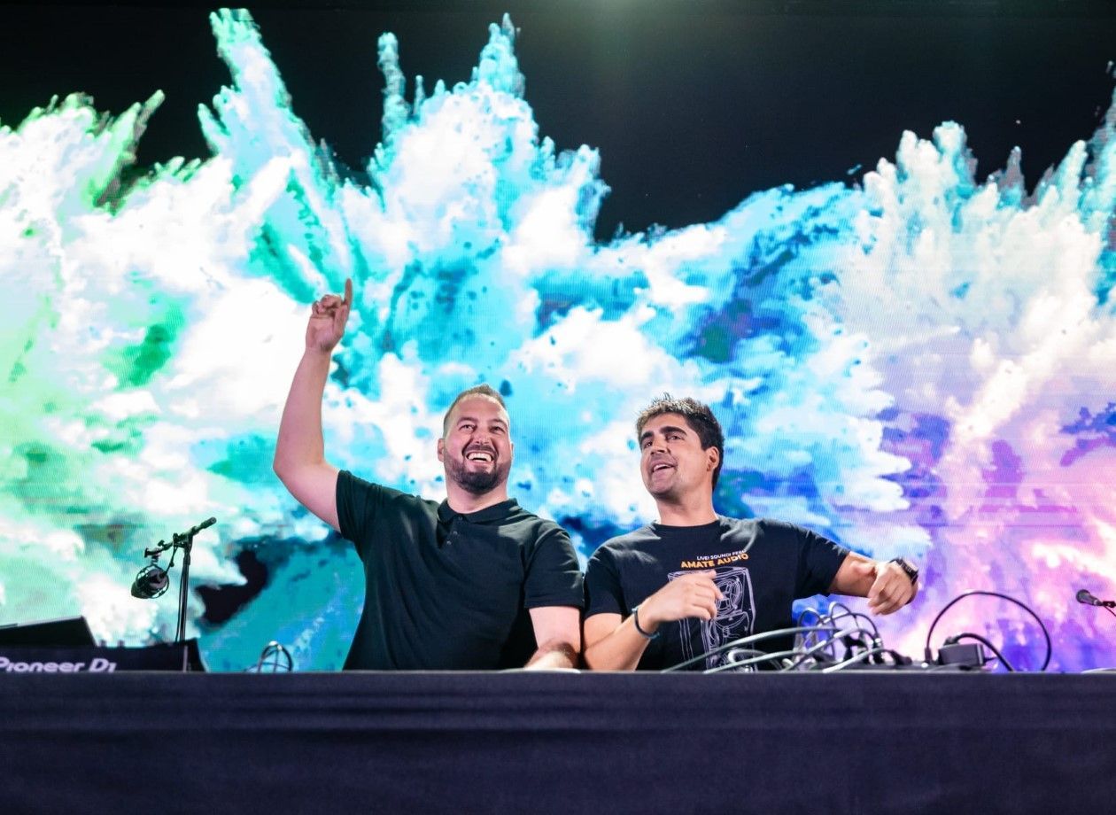 Dos DJs en un concierto, sonriendo ante un fondo abstracto y colorido. Uno levanta el brazo.