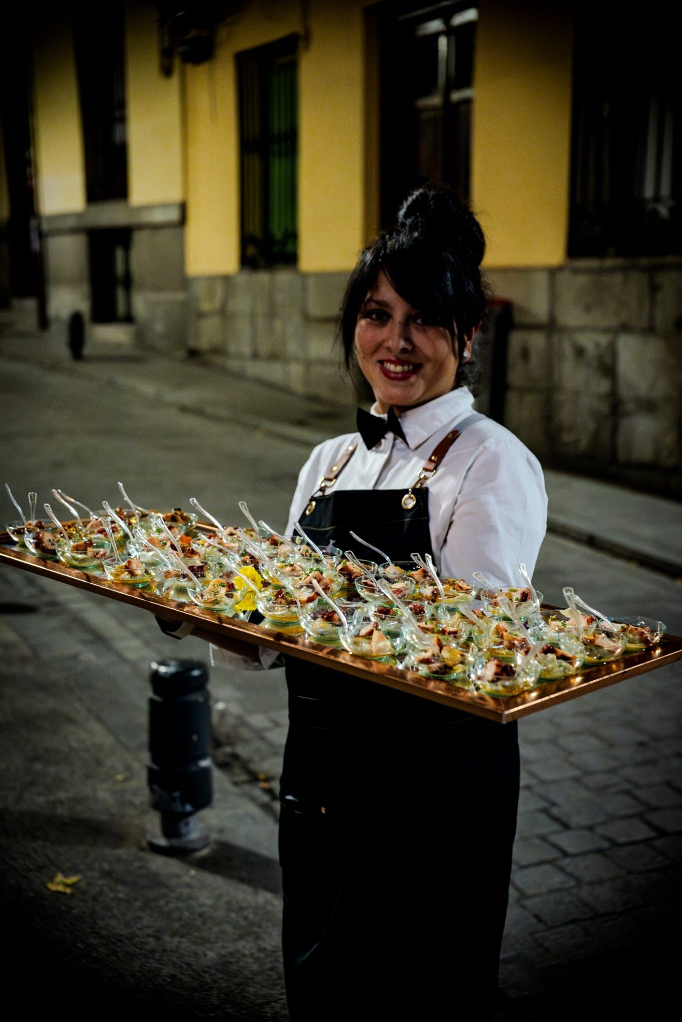 Un camarero sonriente con uniforme formal sostiene una bandeja de aperitivos en el exterior de un edificio al anochecer.