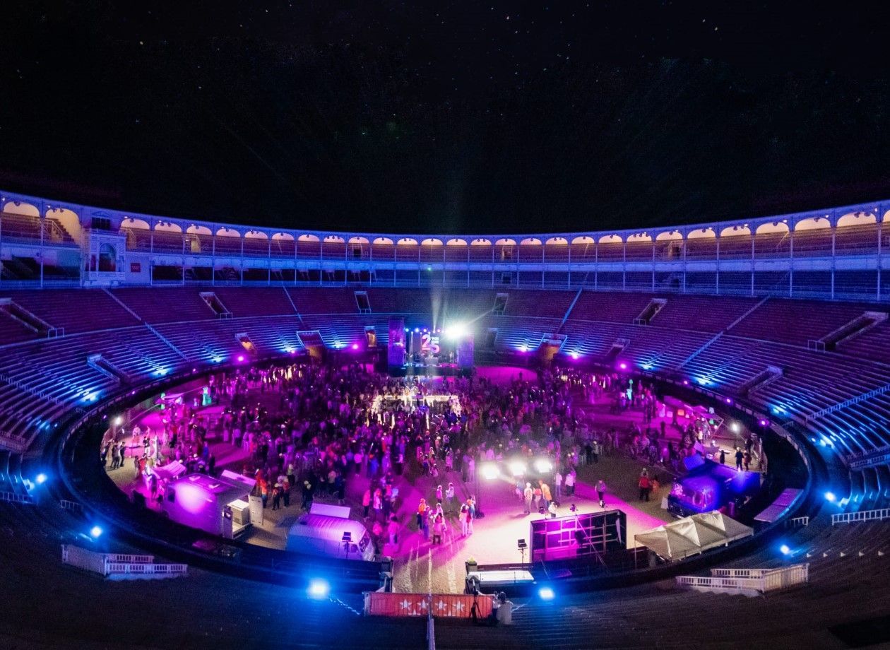 Vista nocturna de una gran multitud reunida en una plaza de toros, iluminada por luces de escenario de color púrpura y azul, con un escenario instalado en el centro.