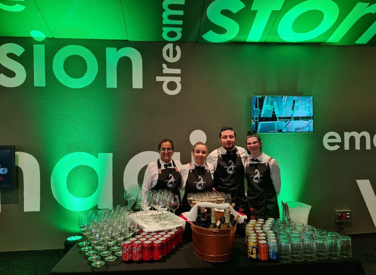 Cuatro camareros con delantales y chalecos están de pie detrás de una mesa llena de bebidas, con 