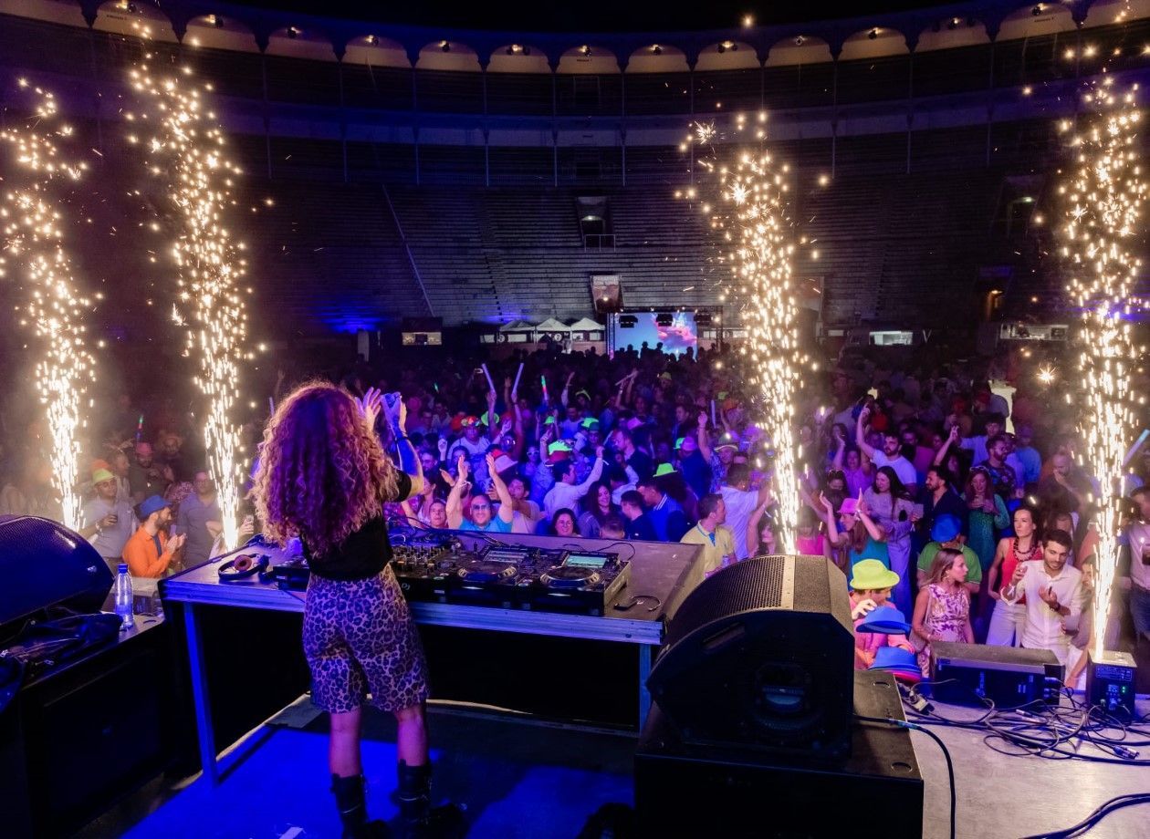 DJ tocando en el escenario, desprendiendo chispas. Público enérgico con las manos en alto. El local iluminado con luces azules y moradas.