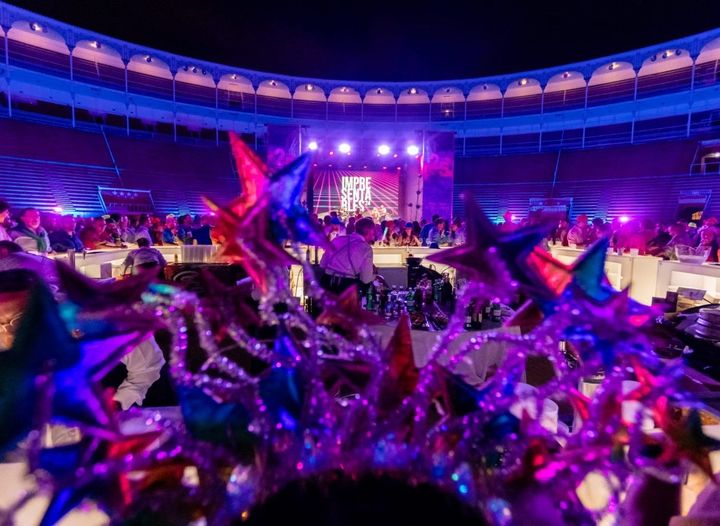 Un evento nocturno en un gran estadio, mesas preparadas para una multitud, escenario iluminado con morado y azul, decoraciones de estrellas en primer plano.