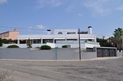 Casa moderna blanca detrás de un muro bajo gris en una calle tranquila bajo un cielo azul.