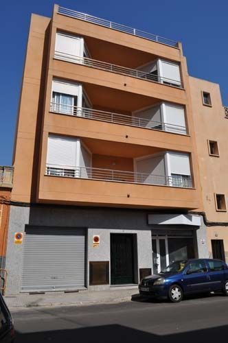 Edificio de apartamentos naranja con balcones y coches aparcados en una calle soleada.