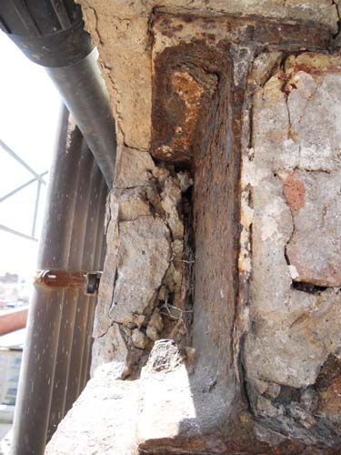 Primer plano de un muro de hormigón dañado con barras de refuerzo expuestas y yeso agrietado junto a un poste metálico.