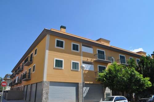Edificio de apartamentos en esquina con fachada color mostaza y blanca, balcones y coches aparcados bajo un cielo azul despejado.