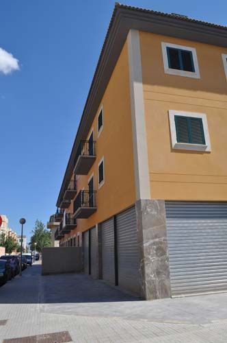 Edificio de apartamentos con fachada naranja, contraventanas metálicas cerradas y una acera estrecha bajo un cielo azul.