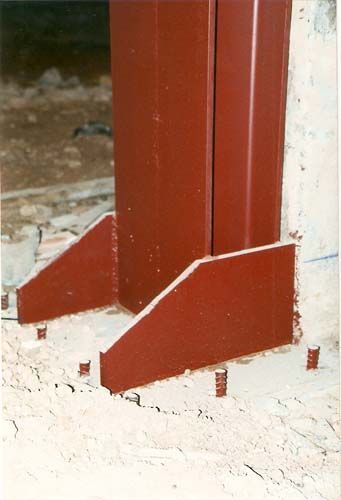 Base de columna de acero rojo con placas de refuerzo triangulares sobre un suelo blanco.