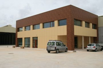 Edificio moderno de dos plantas de color beige y marrón con furgonetas aparcadas delante, en un terreno pavimentado.