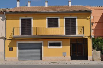 Casa amarilla de dos plantas con balcón, contraventanas oscuras, puerta de garaje y puerta de entrada negra.