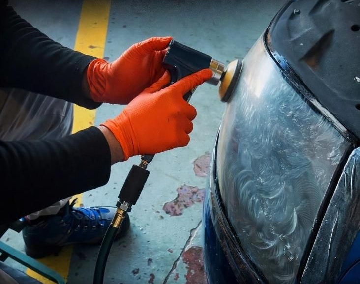 Manos enguantadas lijando el parachoques de un coche con una lijadora eléctrica en un taller.