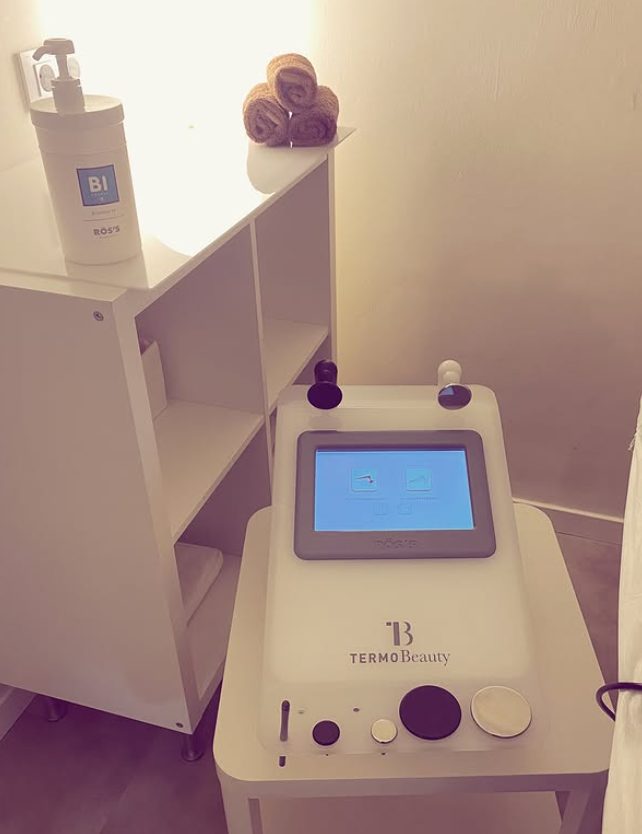 En una pequeña habitación, junto a una estantería blanca y toallas, hay una máquina médica blanca con pantalla táctil.