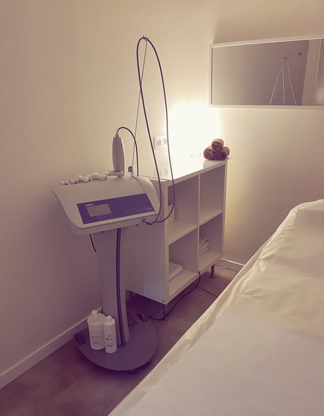 Habitación de clínica con una máquina de tratamiento blanca junto a una cama y un espejo iluminado en la pared.