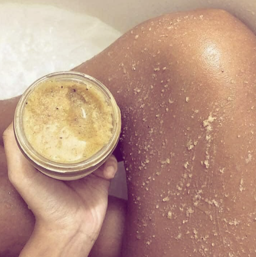 Una persona sostiene un frasco de exfoliante cerca de una pierna cubierta de motas blancas, en un baño o spa.