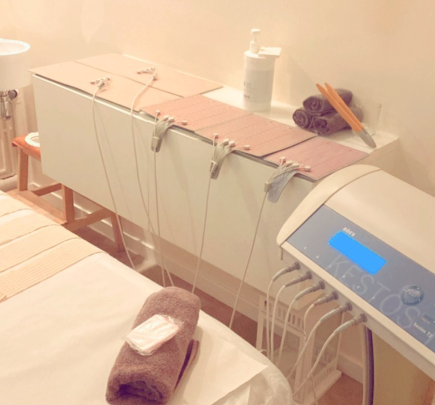Camilla de spa con panel de control azul, toallas, botellas y cables en una habitación con iluminación tenue.