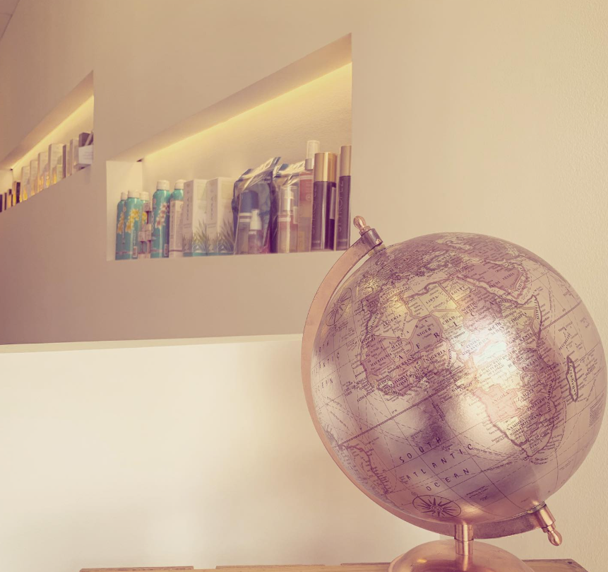 Habitación de tonos cálidos con un globo terráqueo plateado sobre un pedestal y estantes en la pared con libros y botellas.