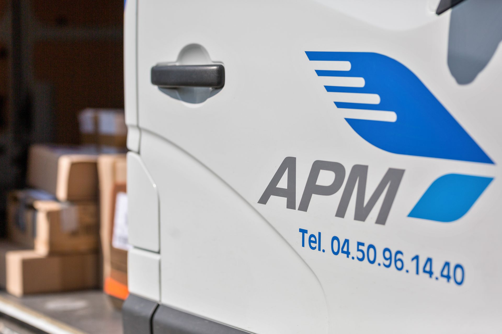 Camionnette de livraison APM blanche avec logo bleu et numéro de téléphone sur le côté, garée près de cartons empilés