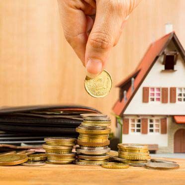 Mano dejando caer una moneda sobre una pila. Al fondo, una casa en miniatura. También se ven una billetera y otras monedas.