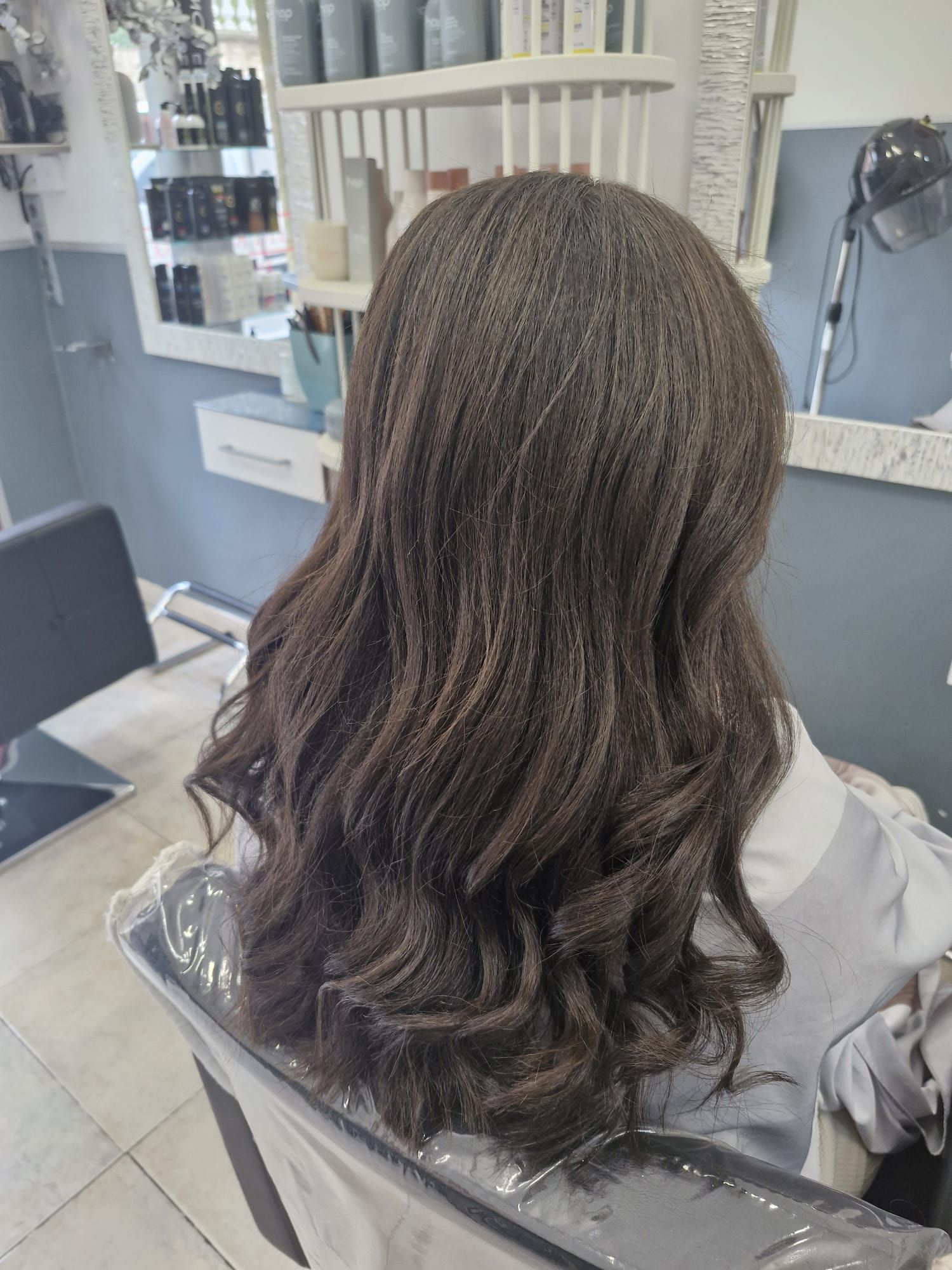 Persona en silla de peluquería con cabello castaño oscuro y ondulado, peinado. Salón con espejo y productos visibles.