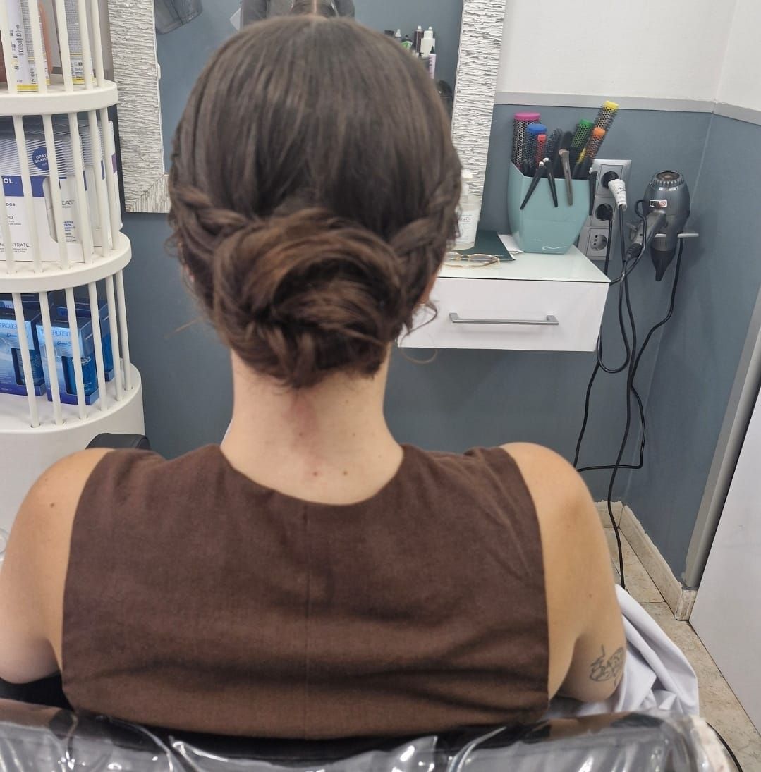 Mujer con cabello oscuro recogido en un moño. Top marrón, sentada en una silla de peluquería. Herramientas de peluquería al fondo.