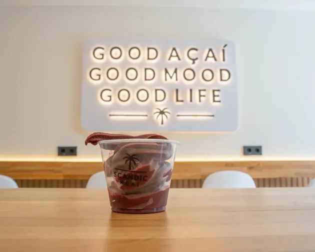 Bebida helada en un vaso transparente sobre una mesa de madera en una cafetería luminosa con un letrero de neón.