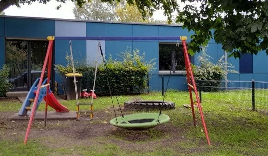 ein Spielplatz mit Schaukeln und Rutsche vor einem blauen Gebäude - Spielplatzprüfung Franziska Müller aus Lüchow im Wendland