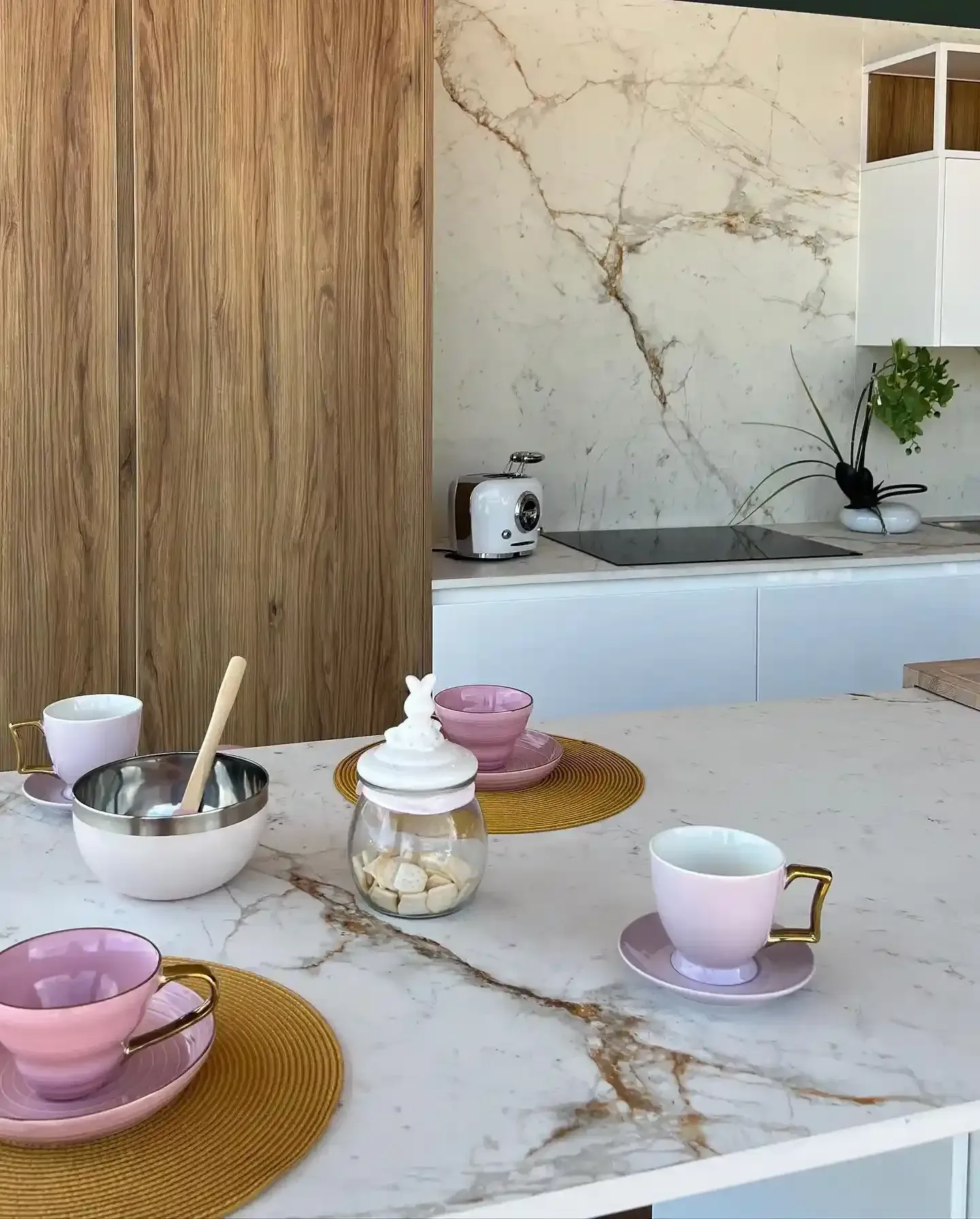 Juego de té en tonos pastel sobre una mesa de cocina de mármol con tazas y cuencos rosas, y un tarro de cristal con galletas.