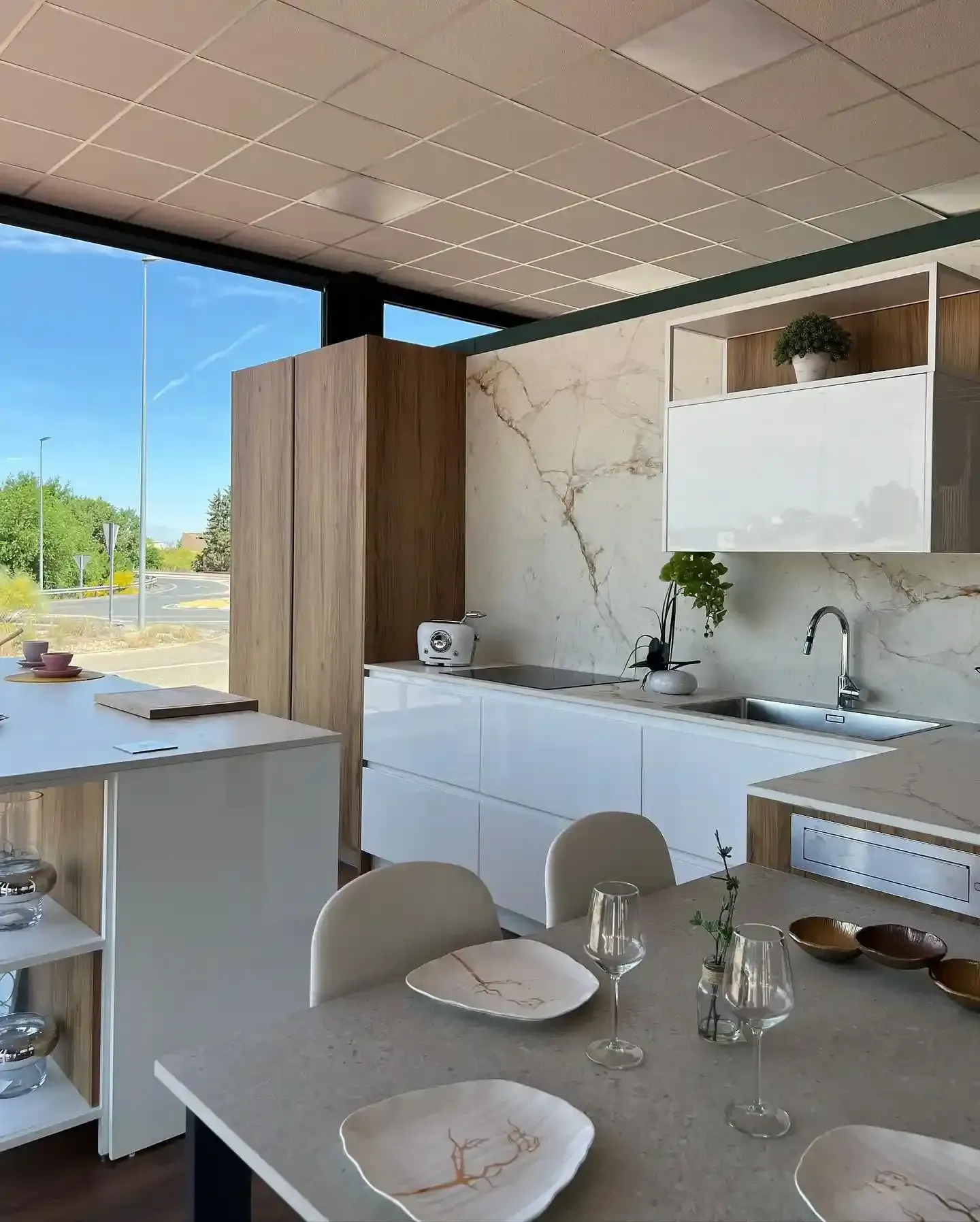 Cocina y comedor modernos con armarios blancos, detalles en madera, salpicadero de mármol y una mesa para dos personas.